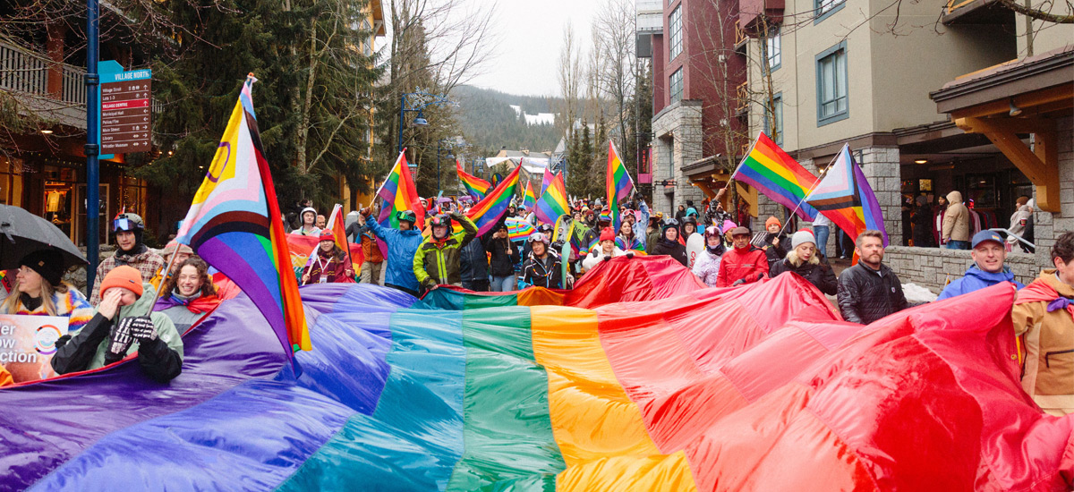 Large Pride parade in Whistler Village with a diverse crowd carrying a wide rainbow banner and waving Pride flags along a pedestrian street lined with shops and mountains in the background.
