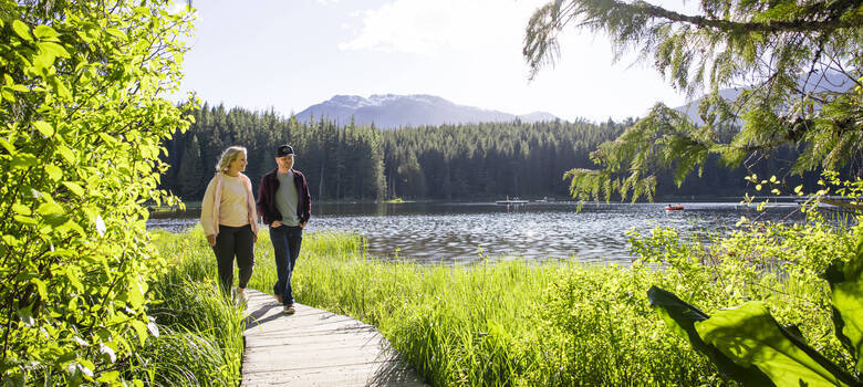 Couple walking on a lakeside boardwalk trail in Whistler, surrounded by lush greenery and mountain views on a sunny day.