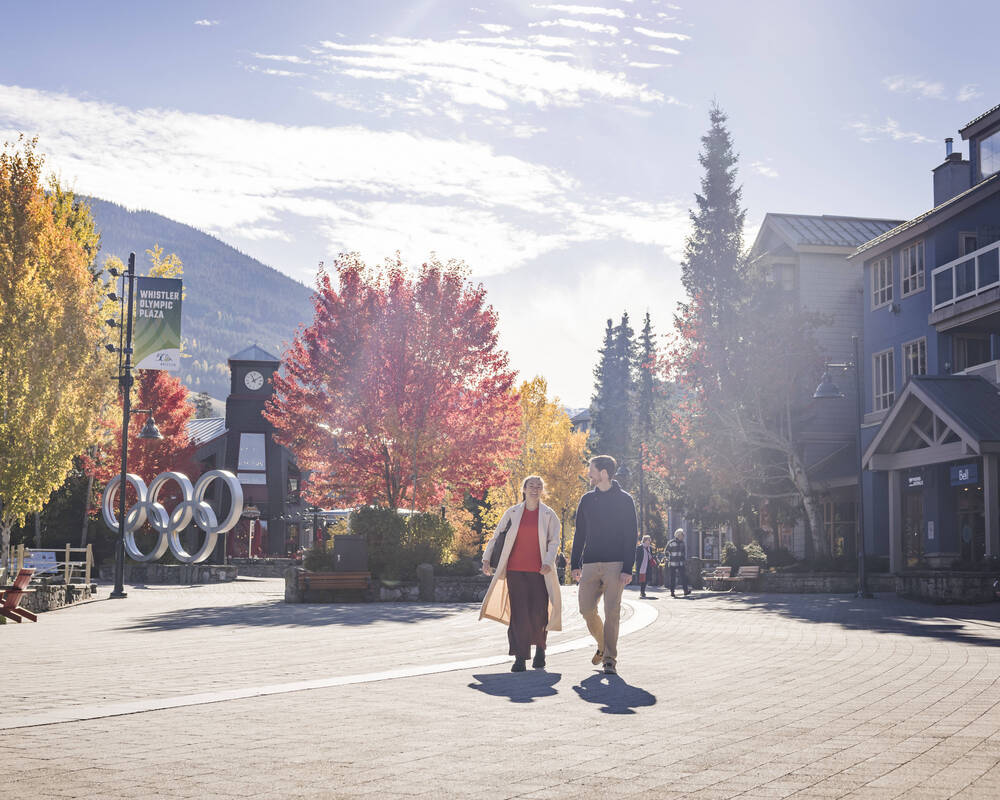 A couple walking along the Whistler Village Stroll in fall with colourful trees and mountains behind them.