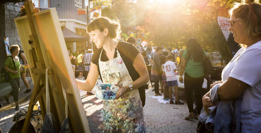 An artist painting outdoors in Whistler Village during golden hour, chatting with a passerby as people enjoy the lively street scene in the background.
