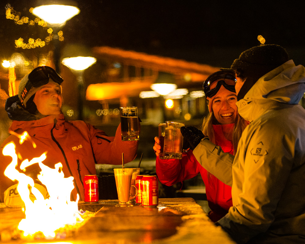 Three friends in ski jackets raise drinks around an outdoor fire table at night