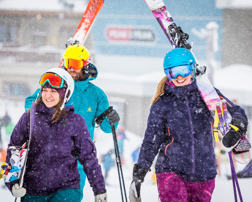 Three smiling skiers in helmets and goggles carry their skis through falling snow
