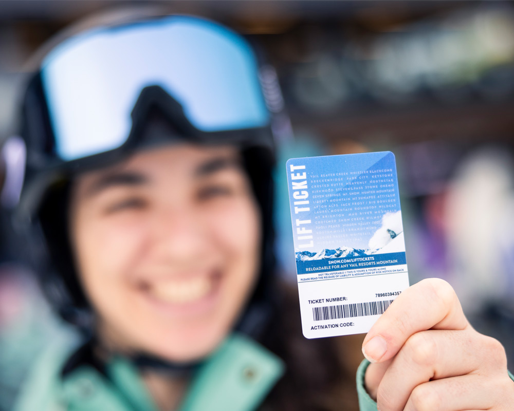 A smiling skier in a helmet and goggles holds up a lift ticket