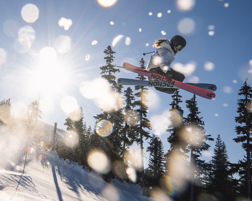 A skier crosses their skis mid-air off a terrain park jump on a sunny day