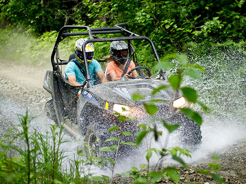 Two riders in a side-by-side UTV speed through a muddy forest trail, water spraying up as they drive through a puddle surrounded by dense greenery.