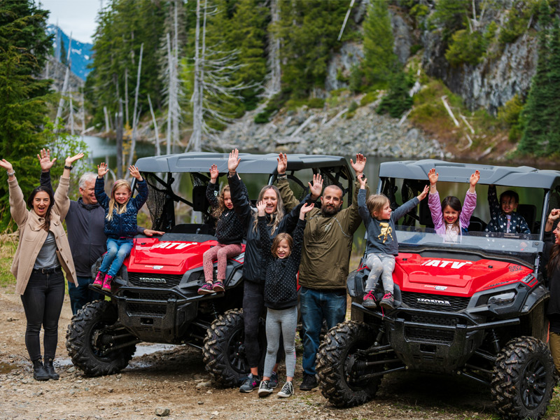 Group of adults and children pose beside two red ATVs on a mountain trail, raising their hands with forested slopes and rocky terrain behind.
