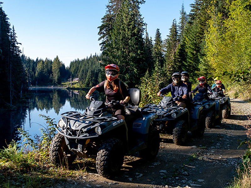 Riders on ATVs follow a lakeside trail through a forest, with calm water reflecting trees and mountains alongside the path.