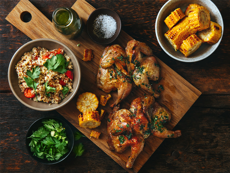 Grilled chicken served on a wooden board with roasted corn, quinoa salad and fresh herbs, arranged on a rustic table.