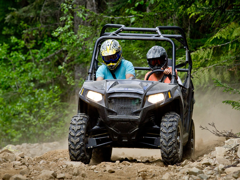 Two riders in a side-by-side UTV drive over a rocky trail in a forest, kicking up dust as they navigate uneven terrain.