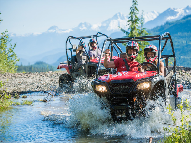 Side-by-side UTVs drive through a shallow creek, splashing water as riders in helmets navigate a rocky alpine trail with mountain views behind.
