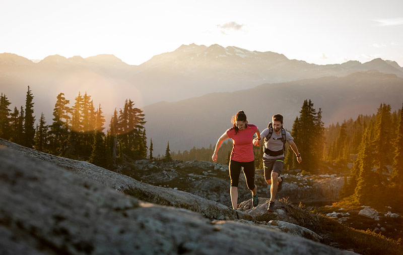 Two hikers make their way up a rocky alpine trail in Whistler at sunset. The golden light casts a warm glow over the rugged mountain landscape, with towering peaks and evergreen trees in the background. They move with energy and determination, fully immersed in the beauty of nature