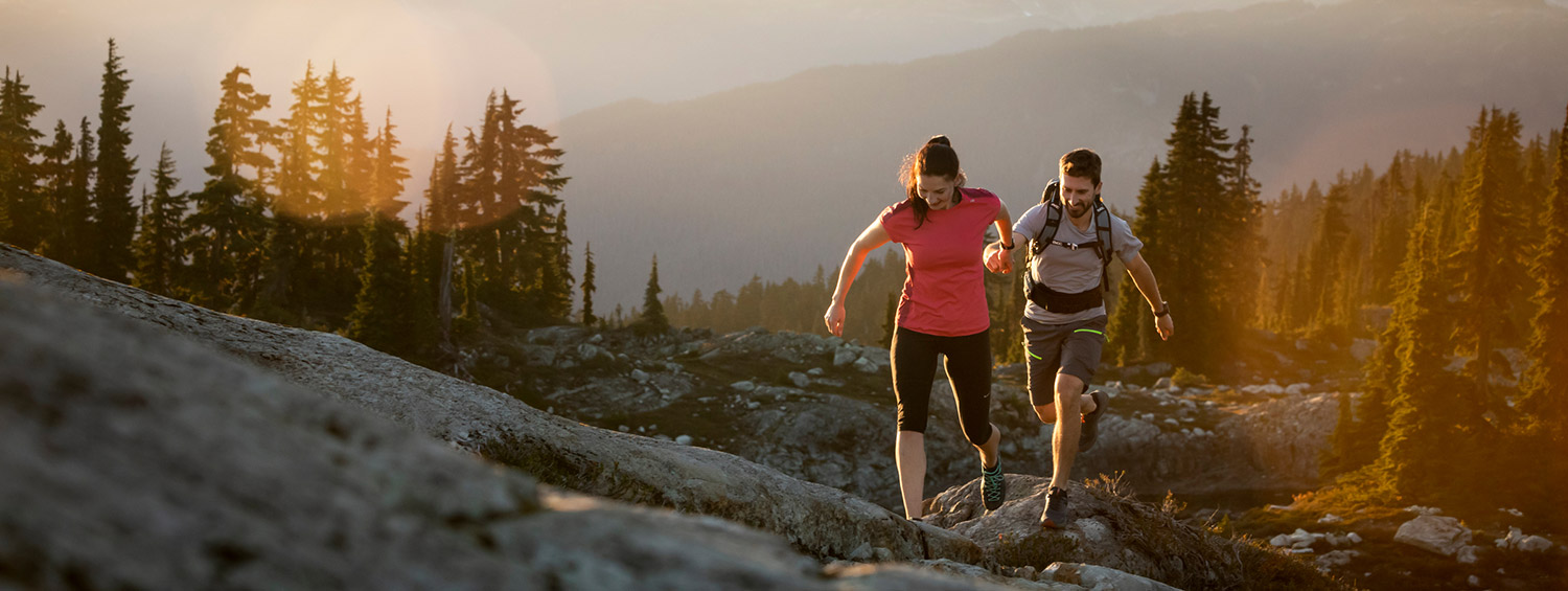 Two hikers make their way up a rocky alpine trail in Whistler at sunset. The golden light casts a warm glow over the rugged mountain landscape, with towering peaks and evergreen trees in the background. They move with energy and determination, fully immersed in the beauty of nature