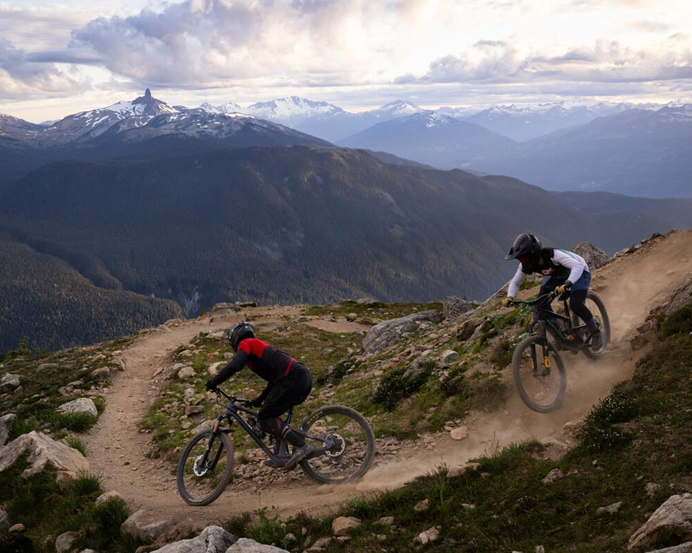 Mountain bikers descend a rugged alpine trail in Whistler Bike Park with sweeping mountain views in the background.