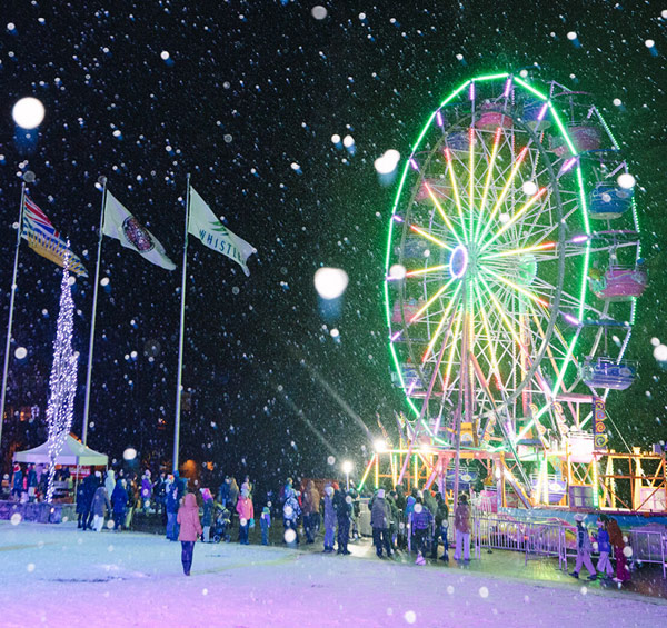 Nighttime winter festival in Whistler with a brightly lit Ferris wheel, falling snow, and people enjoying the festive atmosphere.