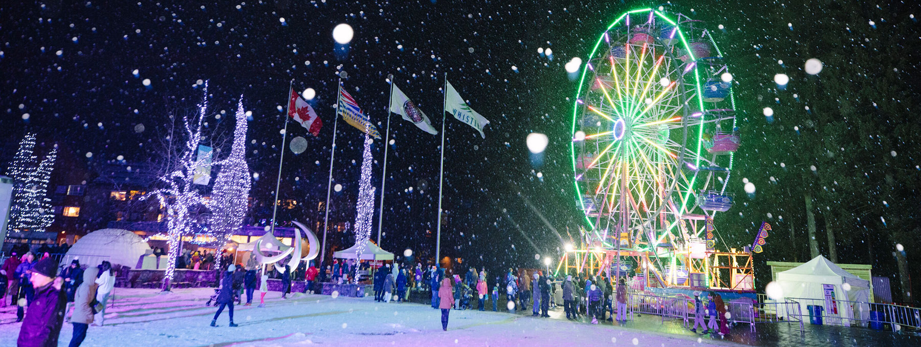 Nighttime winter festival in Whistler with a brightly lit Ferris wheel, falling snow, and people enjoying the festive atmosphere.