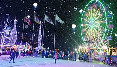 Nighttime winter festival in Whistler with a brightly lit Ferris wheel, falling snow, and people enjoying the festive atmosphere.