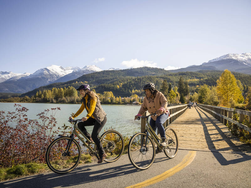 Two people riding cruiser bikes beside Green Lake in Whistler on a sunny day in the fall