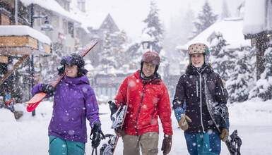 Three people walking with ski and snowboard gear on the snowy Whistler Village Stroll