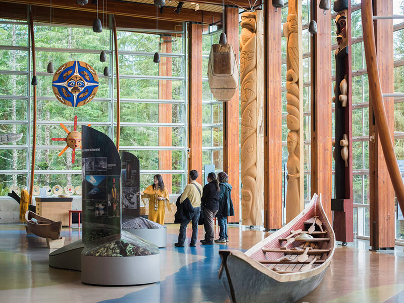 Indigenous cultural ambassador in cedar-bark regalia guiding visitors through the Squamish Lil’wat Cultural Centre gallery in Whistler, BC, featuring hand-carved totem poles, a traditional canoe, and floor-to-ceiling windows overlooking the forest.