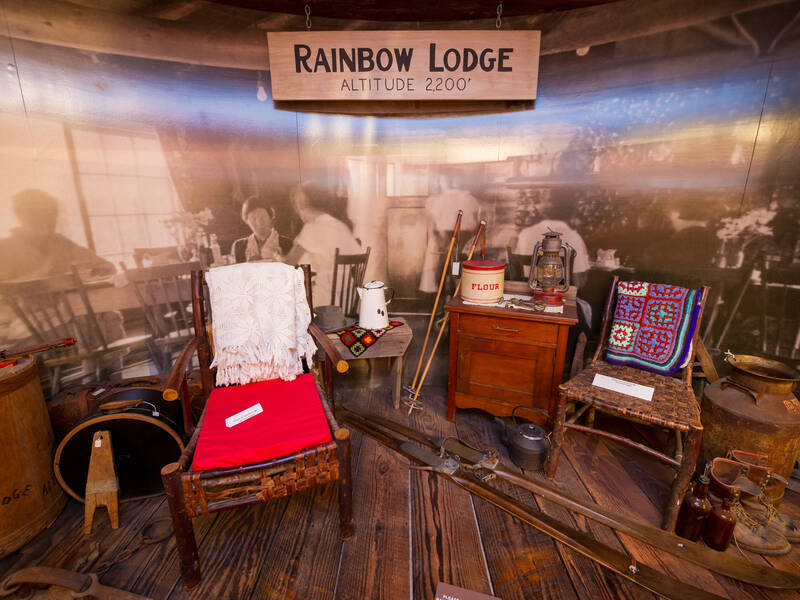 Museum exhibit of Rainbow Lodge artefacts at 2,200 ft in Whistler, BC, featuring a hand-lettered “Rainbow Lodge Altitude 2,200’” sign, rustic wooden chairs draped with crocheted blankets, vintage skis and poles, enamelware coffee pot, lantern and other period pieces.