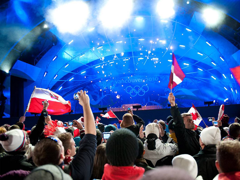 Outdoor Vancouver 2010 Winter Olympics medal ceremony at Whistler Medal Plaza, BC, with spectators waving Canadian and Polish flags under bright stage lights and a blue-lit “Vancouver 2010” stage bearing the Olympic rings.