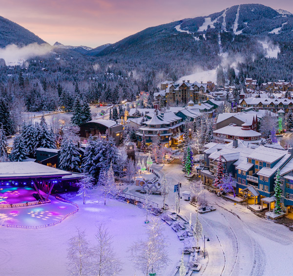 Aerial view of Whistler Village at dusk in winter, with Olympic Plaza's lit stage and Olympic rings below snowy Whistler and Blackcomb mountains
