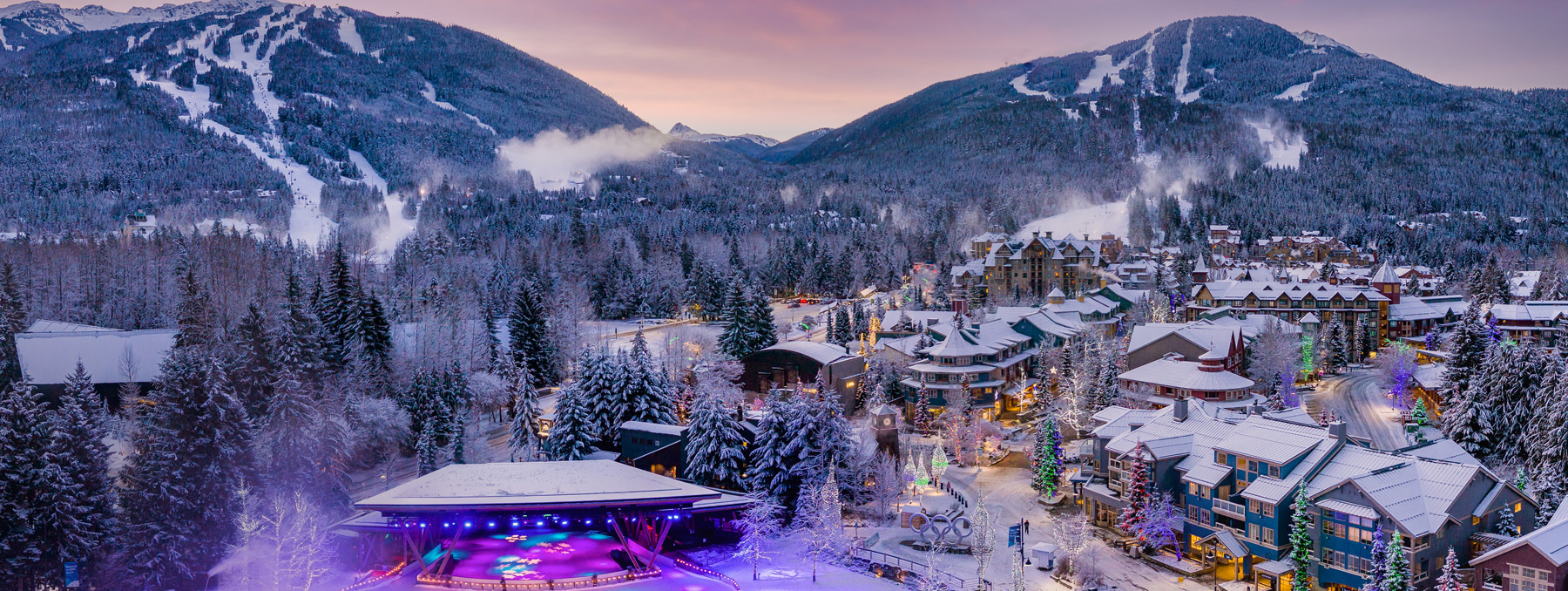Aerial view of Whistler Village at dusk in winter, with Olympic Plaza's lit stage and Olympic rings below snowy Whistler and Blackcomb mountains