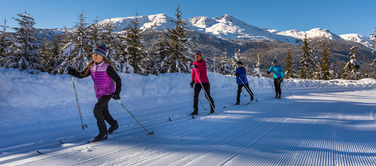 Family cross-country skiing on groomed trails at Whistler Olympic Park under clear winter skies.