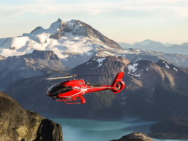 Red helicopter flying above alpine lakes and rugged snow-covered peaks on a scenic helicopter tour near Whistler Blackcomb at sunset.