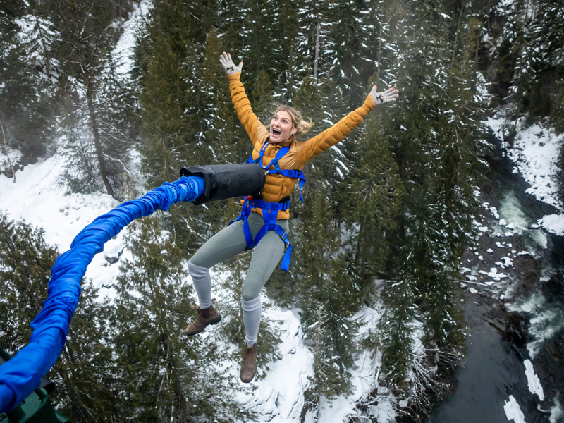 Smiling woman in a yellow sweater and blue harness bungee jumping with arms raised over a snowy evergreen canyon at Whistler Bungee near Whistler, BC, in winter.
