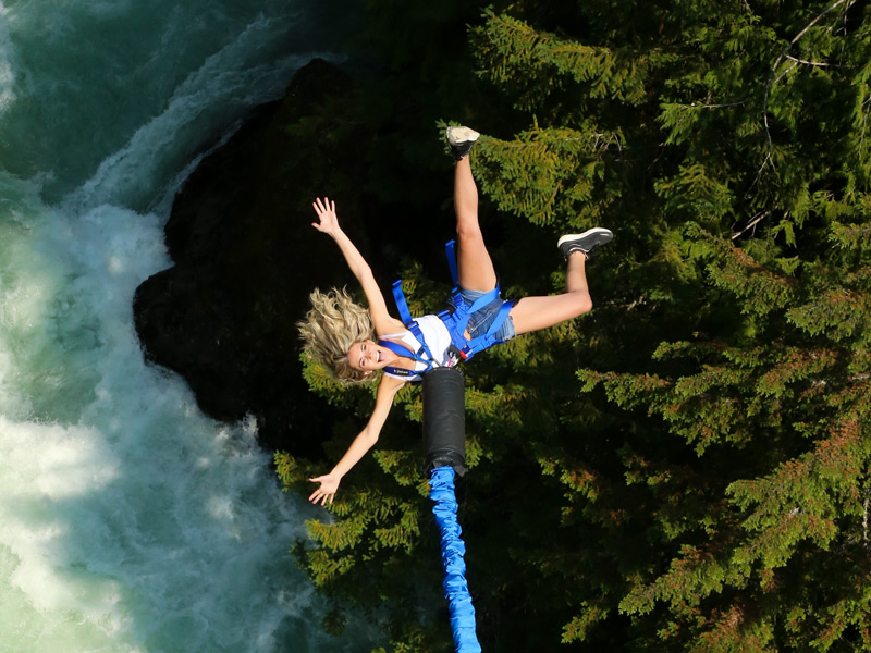 Woman mid-bungee jump in Whistler over a turquoise river, suspended upside down with arms outstretched above a forested canyon.