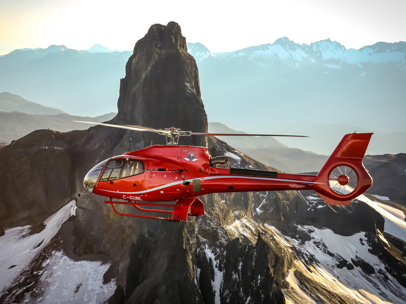 Red helicopter flying past a rugged mountain peak near Whistler Blackcomb during a scenic alpine helicopter tour at golden hour.