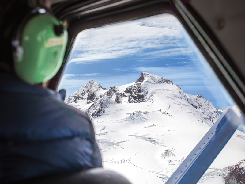 Passenger wearing a headset looks out from a helicopter at a snow-covered glacier and rugged mountain peaks on a scenic flight near Whistler.