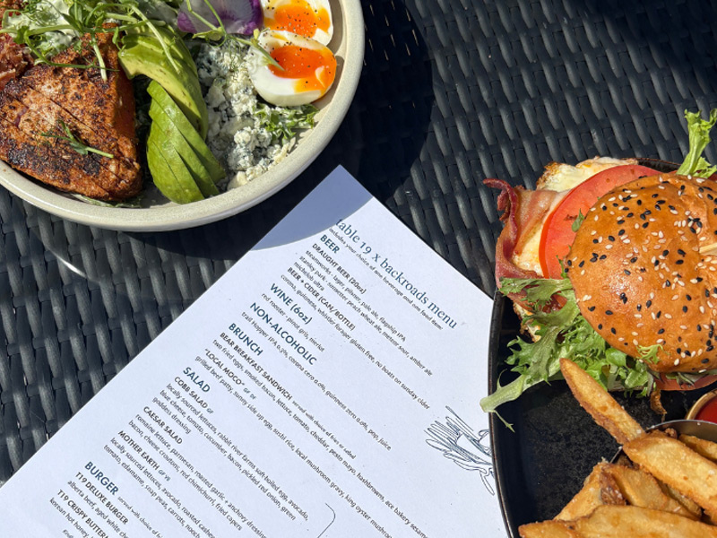 Brunch spread on a sunny Whistler restaurant patio featuring a gourmet burger with fries, a salmon salad with soft-boiled eggs, and an outdoor dining menu.