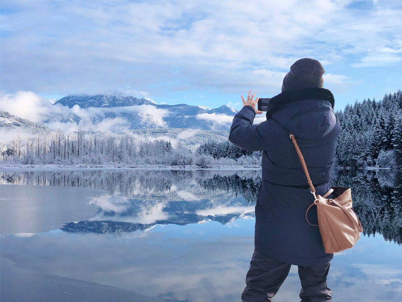A visitor photographs snowy mountains and frosted forest reflected in a calm lake on a crisp winter day in Whistler, BC.