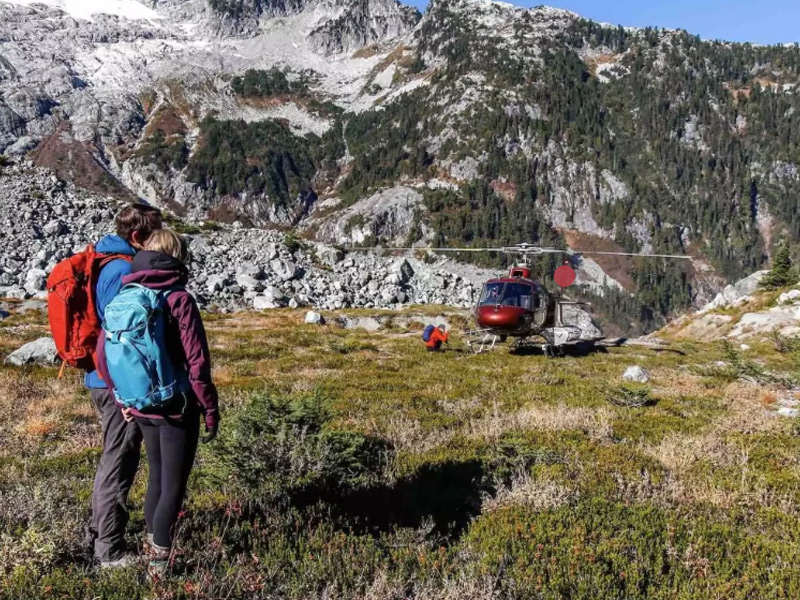 Two hikers with backpacks standing beside a helicopter in an alpine meadow surrounded by rugged mountains during a heli-hiking adventure near Whistler.