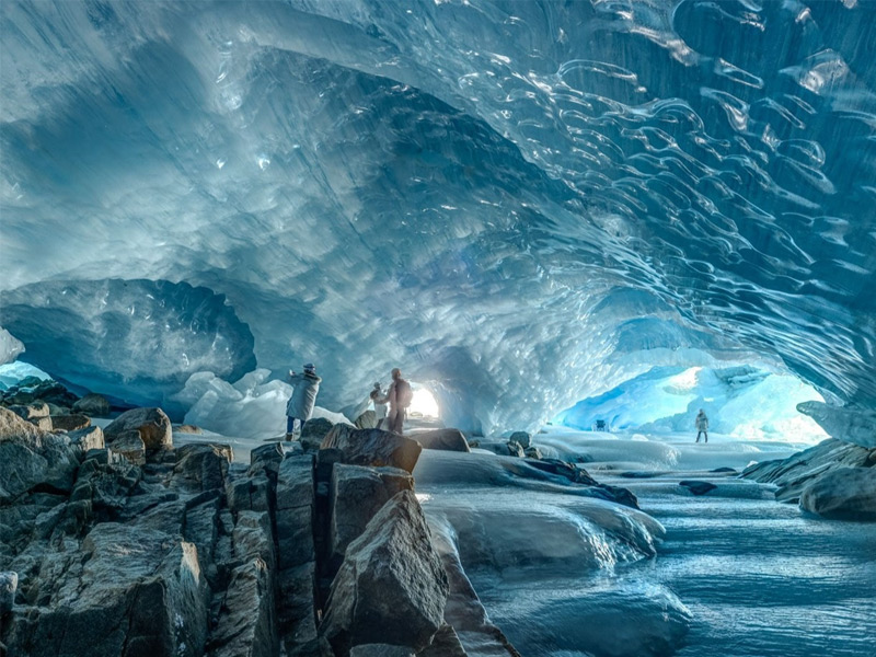 Visitors exploring a glowing blue glacier ice cave surrounded by frozen walls and icy streams on a helicopter adventure tour near Whistler.
