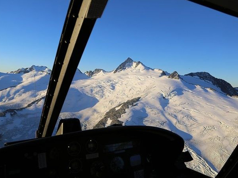View from inside a helicopter flying toward snow-covered alpine peaks and glaciers on a scenic Whistler mountain helicopter tour.