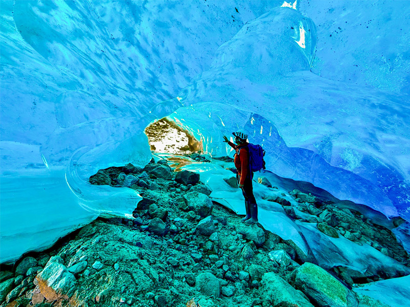 Person standing inside a glowing blue glacier ice cave during a guided alpine adventure near Whistler, surrounded by smooth ice walls and rocky terrain.