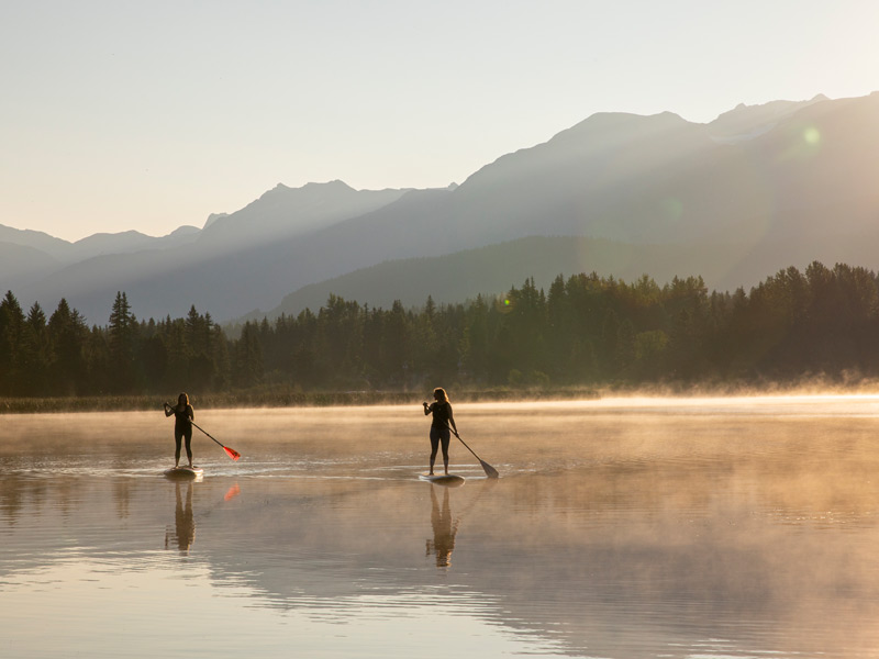People enjoying stand up paddleboards, canoes and kayaks on a calm lake in Whistler.