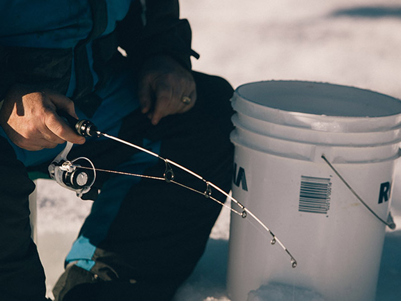 Close-up of an angler's hands holding a short ice fishing rod and reel beside a white bucket on a snowy frozen lake in Whistler.