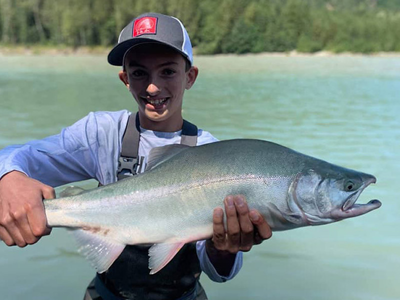 A smiling young angler in a cap holds up a large pink salmon on a glacier-fed river during a summer fishing trip near Whistler, BC.