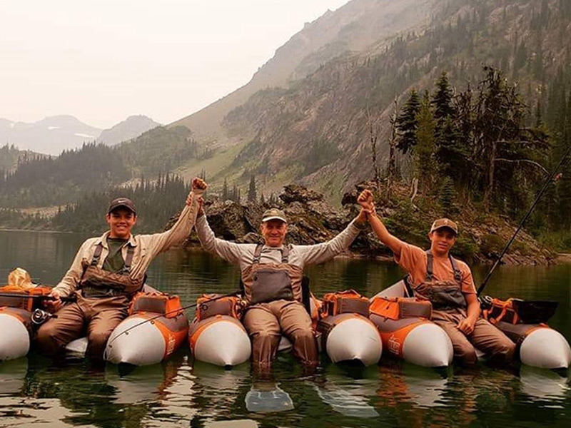 Three anglers in waders raise their joined hands in celebration while float tube fishing on a calm alpine lake below rugged mountains near Whistler.