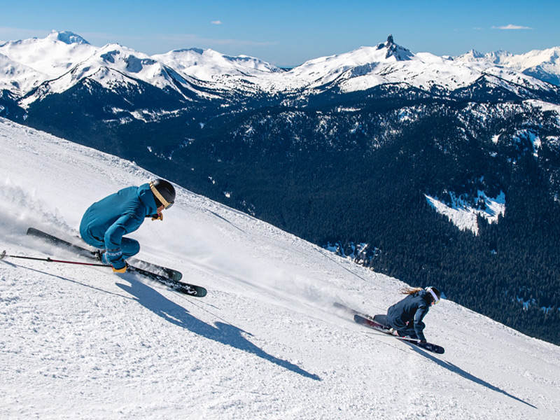 Two skiers carve down a snowy alpine run in Whistler with Black Tusk and the Coast Mountains in the distance