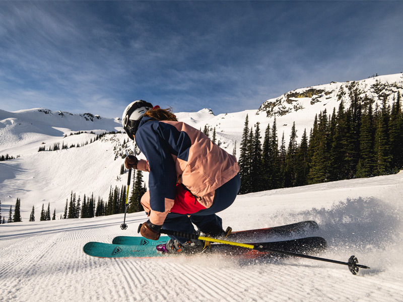 Skier carves across a freshly groomed ski run in Whistler, kicking up snow beneath the surrounding mountains