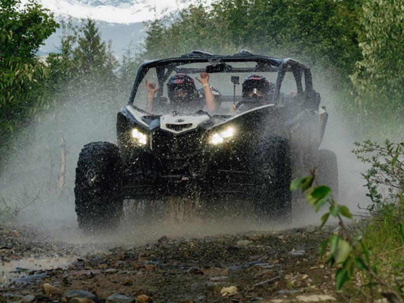 Two riders in a side-by-side UTV speed through a muddy forest trail, water spraying up as they drive through a puddle surrounded by dense greenery.