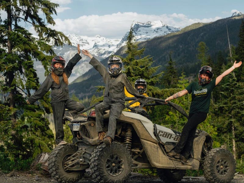 Two riders in a side-by-side UTV drive over a rocky trail in a forest, kicking up dust as they navigate uneven terrain.