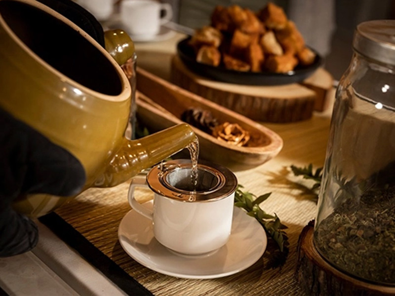 Hot tea is poured through a strainer into a cup beside pastries and local ingredients in a cozy setting.