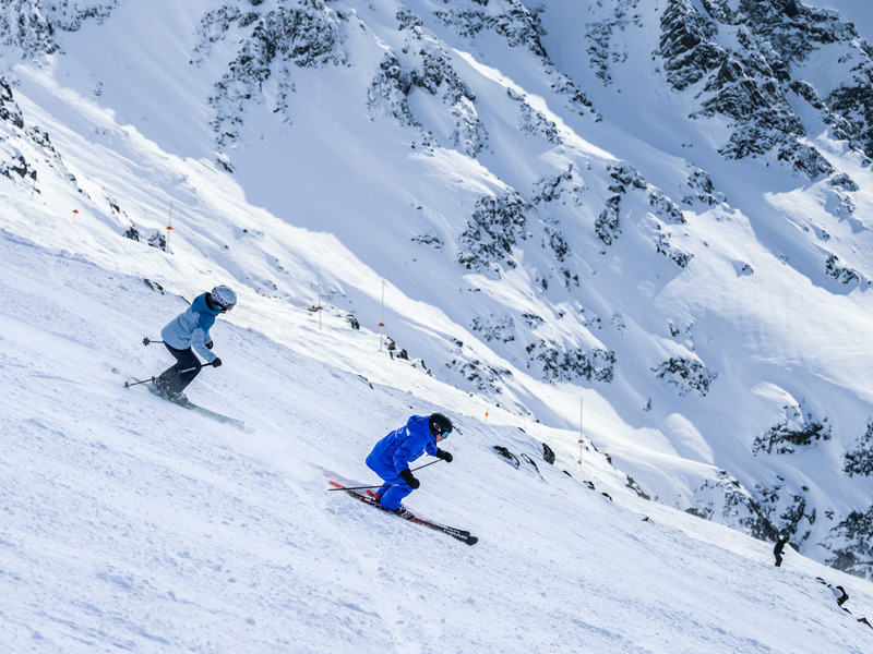 Two skiers carve down a snowy alpine run in Whistler with Black Tusk and the Coast Mountains in the distance