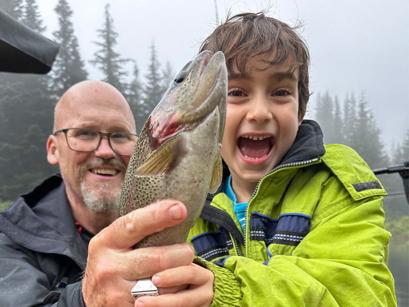 An excited young boy and a smiling guide show off a speckled rainbow trout on a misty lake surrounded by evergreen forest near Whistler.